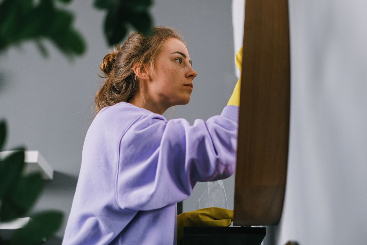 services-04 Vue de profil d’une jeune femme concentrée, portant des gants en latex, debout et nettoyant un miroir dans une pièce lumineuse en journée